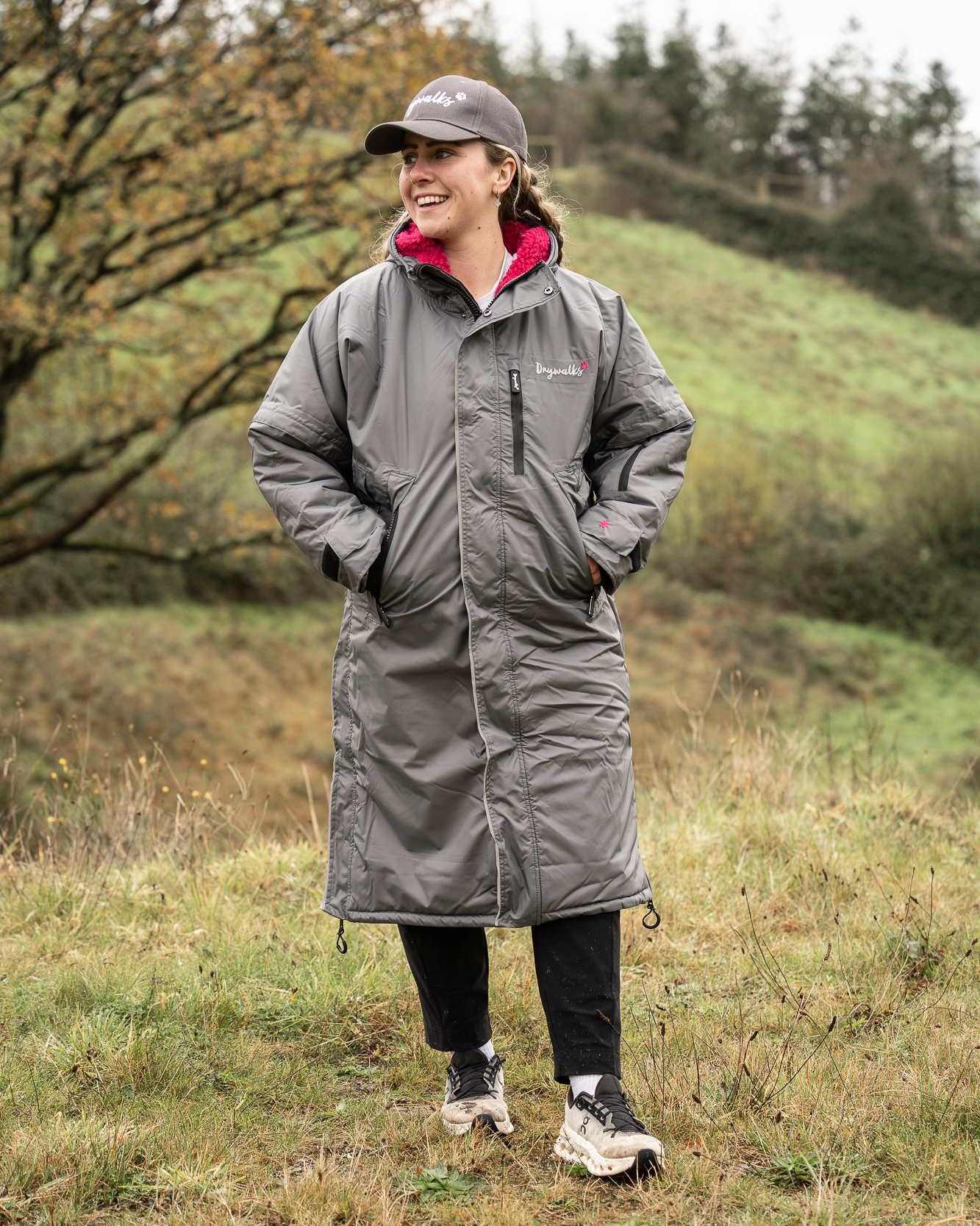 Person wearing a grey long oversized Drywalks coat standing in a grassy field with trees in the background